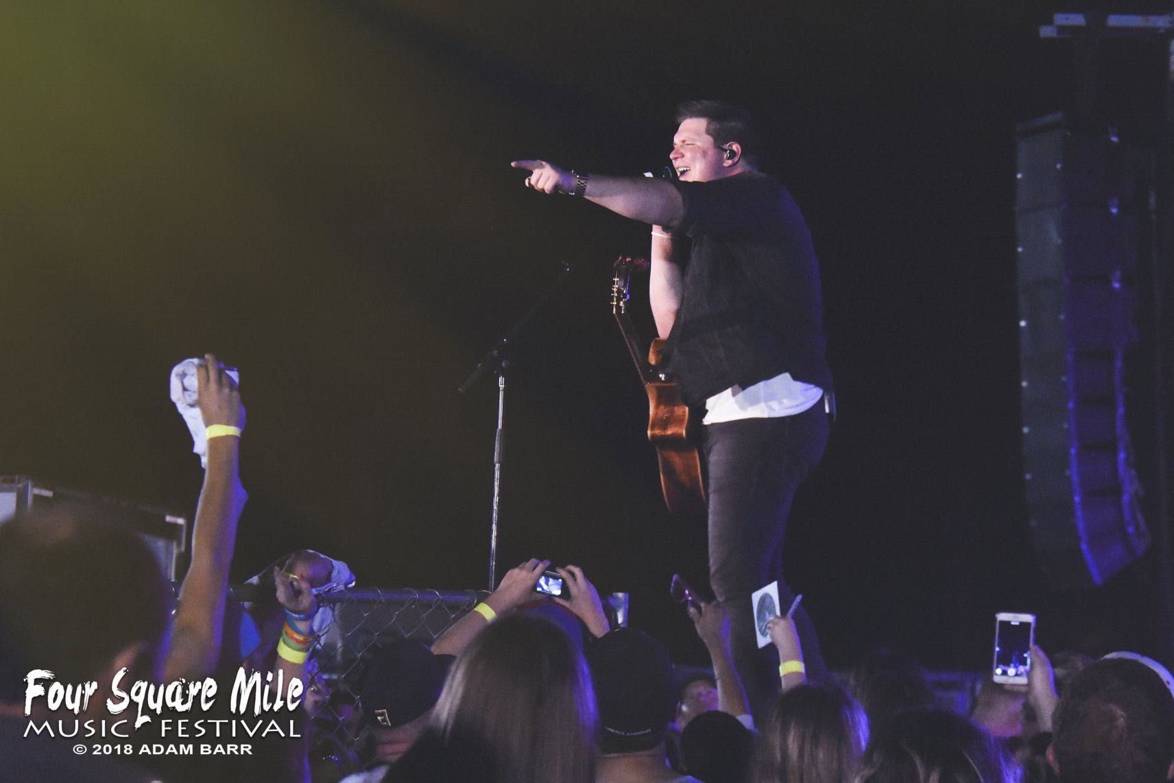 Man on stage pointing, performing at Four Square Music Festival. Crowd cheering with phones up.