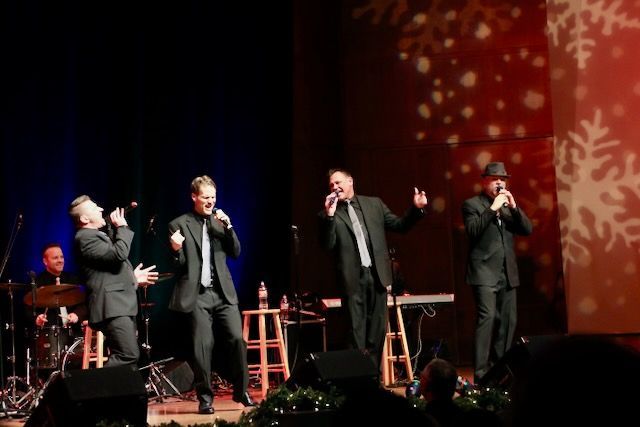 A cappella group on stage, singing into microphones. Red and white snowflake backdrop.
