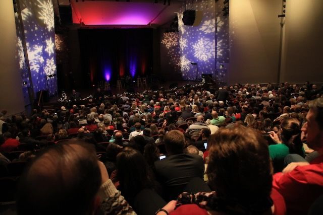 Audience in a theater watches a performance. Stage lit with purple, red, blue lights; snowflakes projected on walls.
