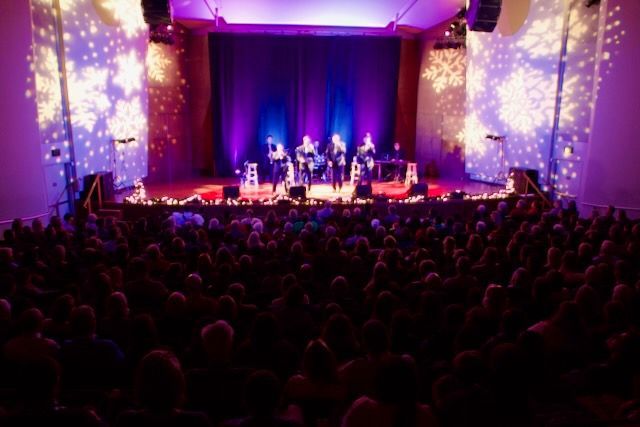 Band performs on stage with audience in a theater. Purple stage and wall lighting, projected snowflakes.