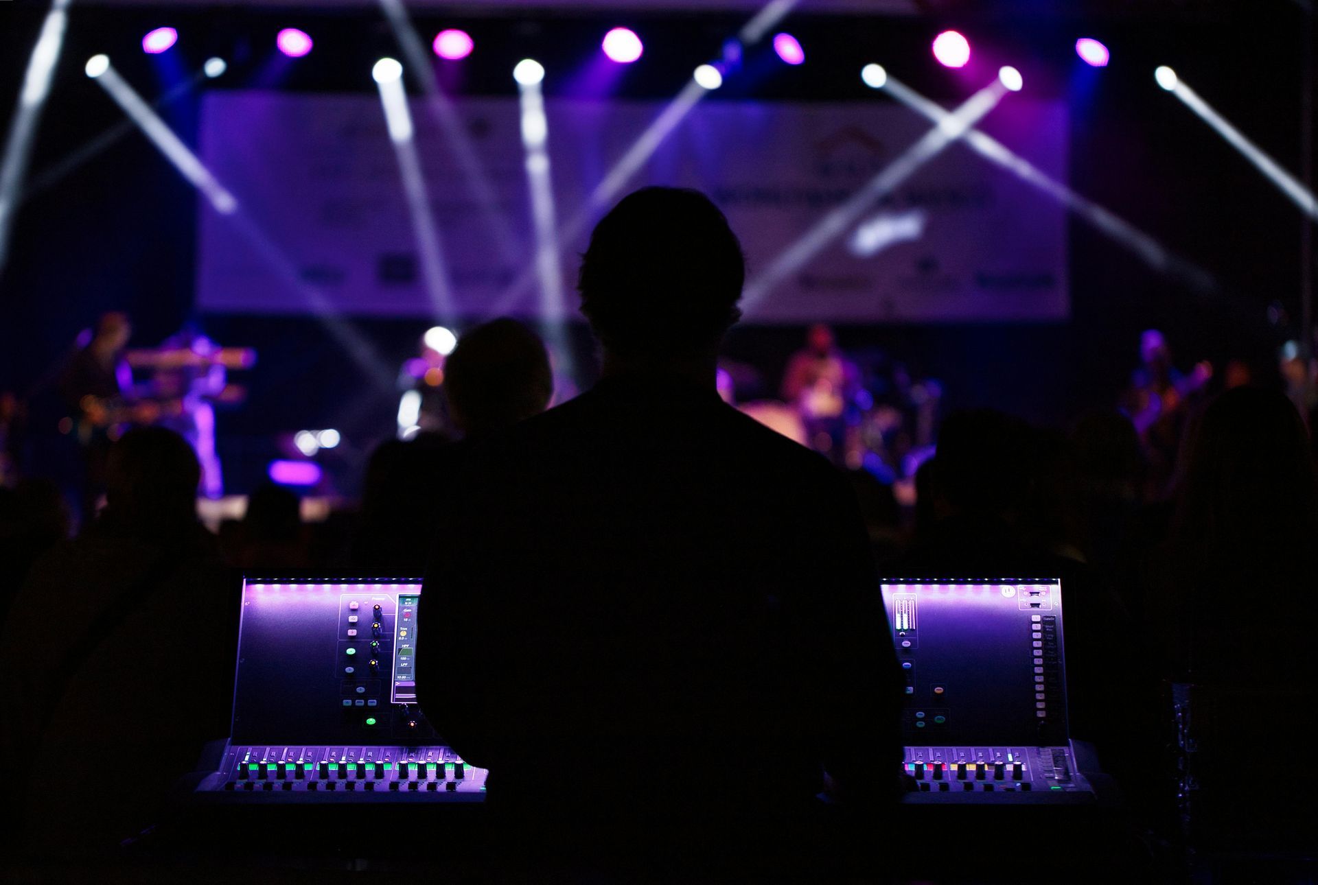 Silhouette of person at sound mixing console, stage with band in background, purple stage lights.