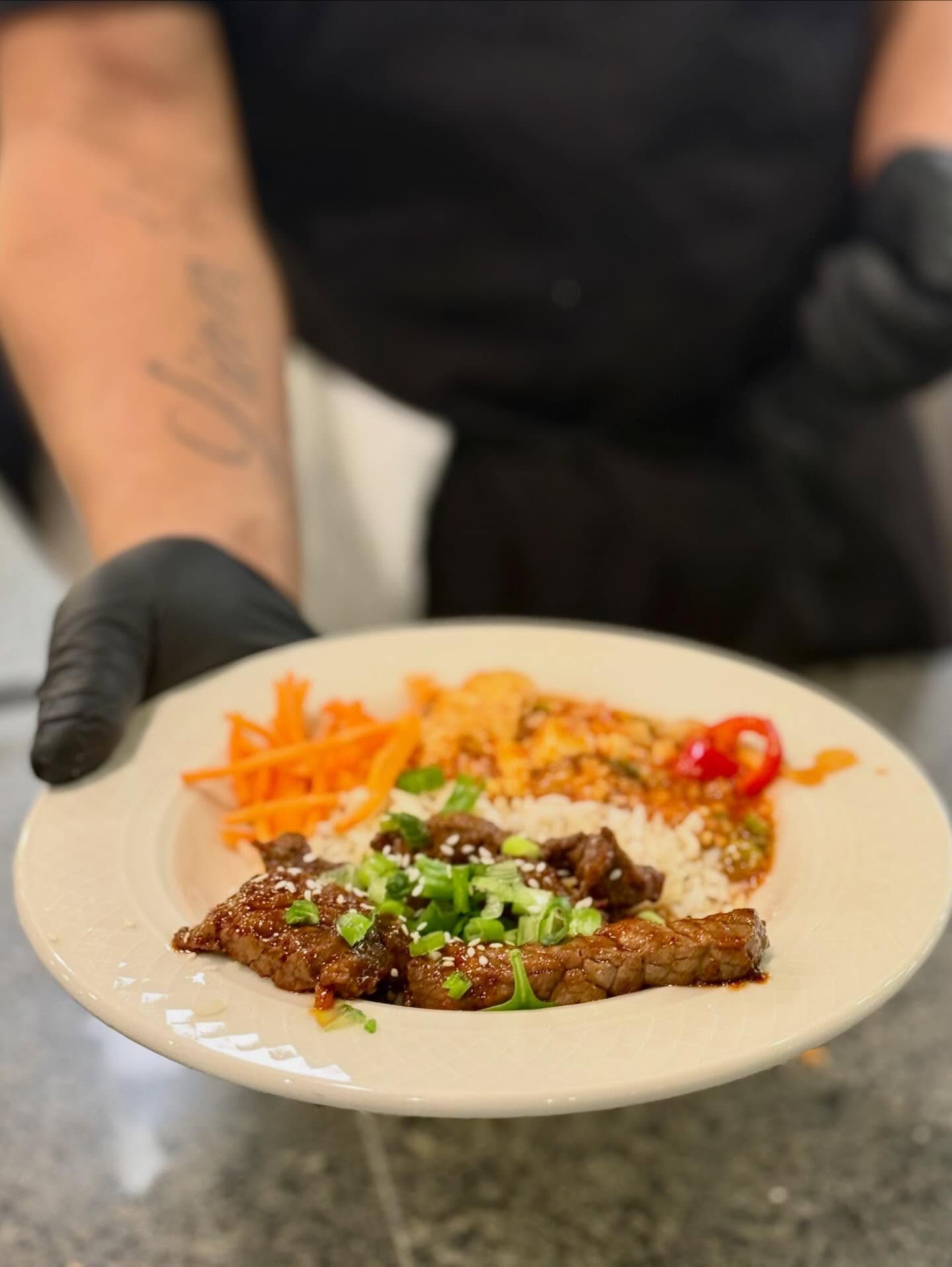 A person in black gloves presents a plate of glazed beef over rice with julienned carrots and garnish.