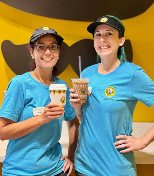 Two smiling workers in blue uniforms and black hats hold beverage cups.