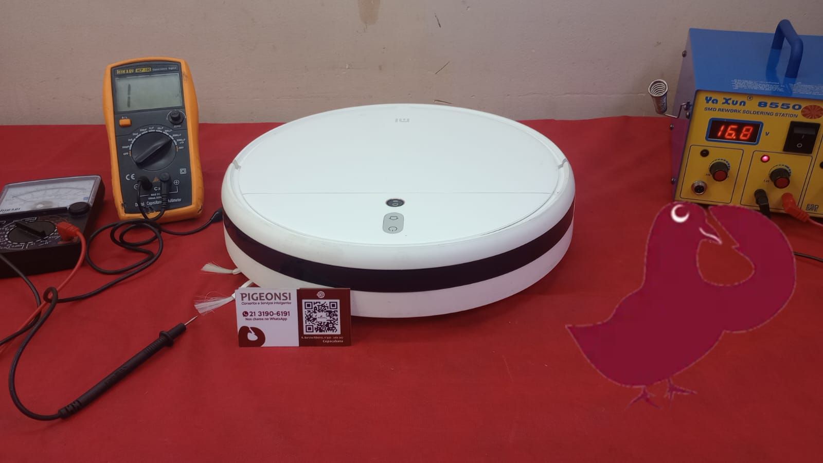 Um aspirador de pó robótico está sobre uma mesa vermelha ao lado de um multímetro.