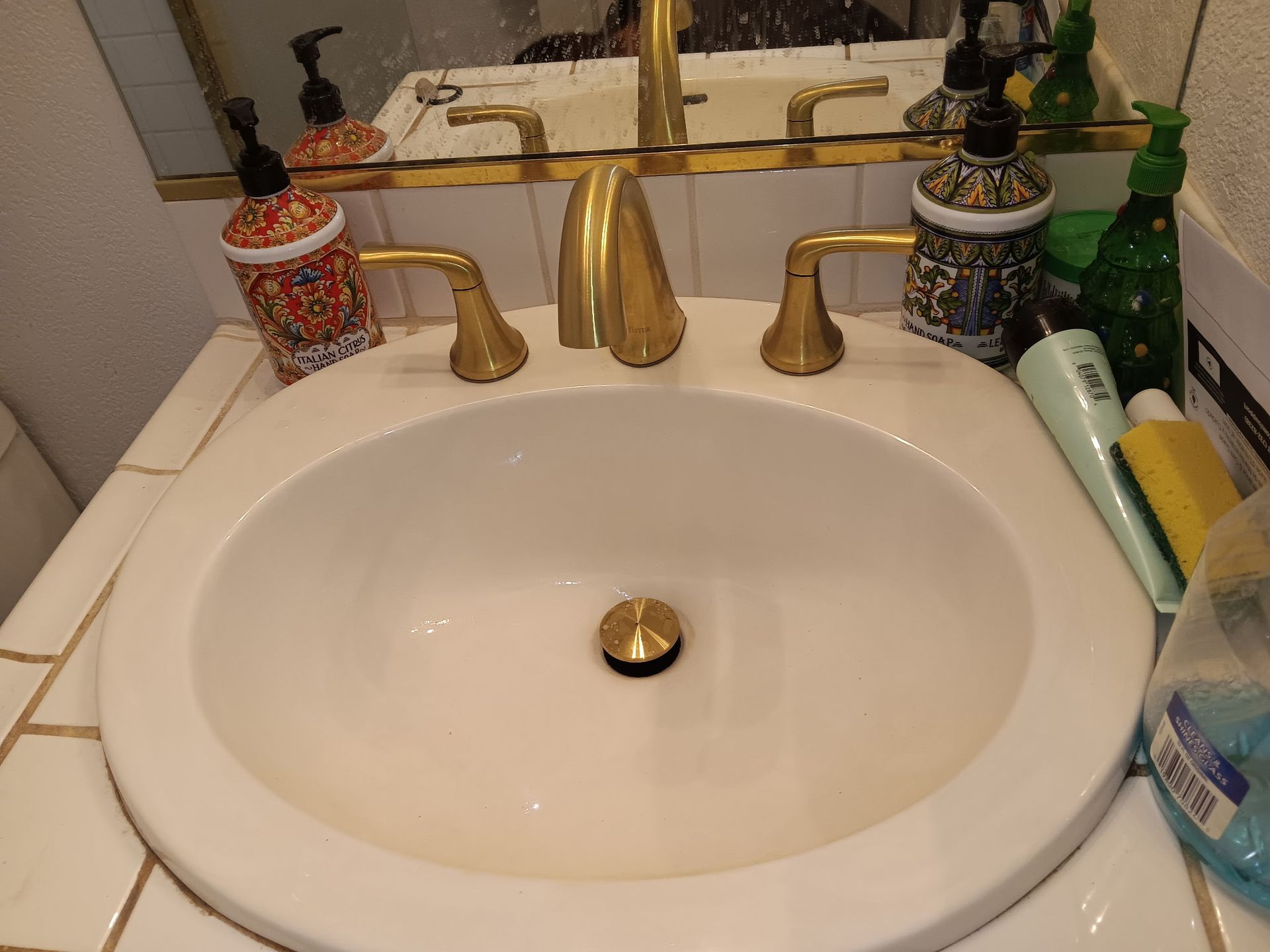Bathroom sink with gold faucet and soap dispensers on a white counter, seen in a mirror.