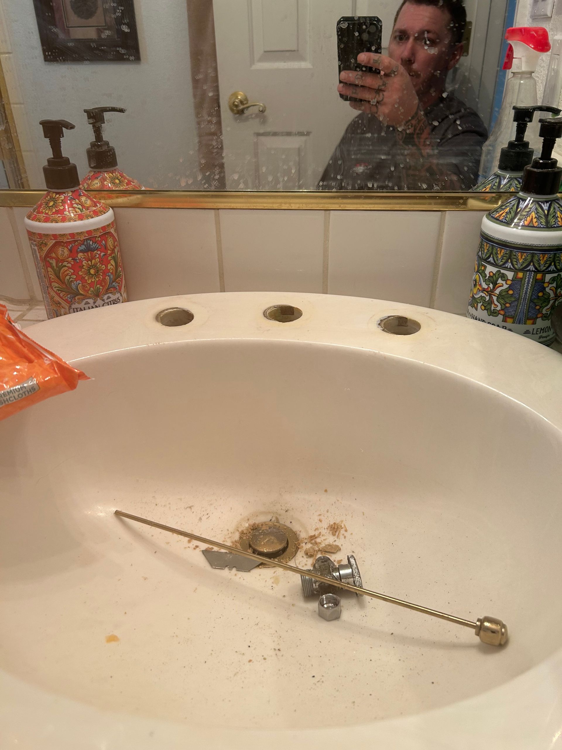 Bathroom sink with a broken metal chain and debris on the basin; a person’s reflection appears in the mirror.