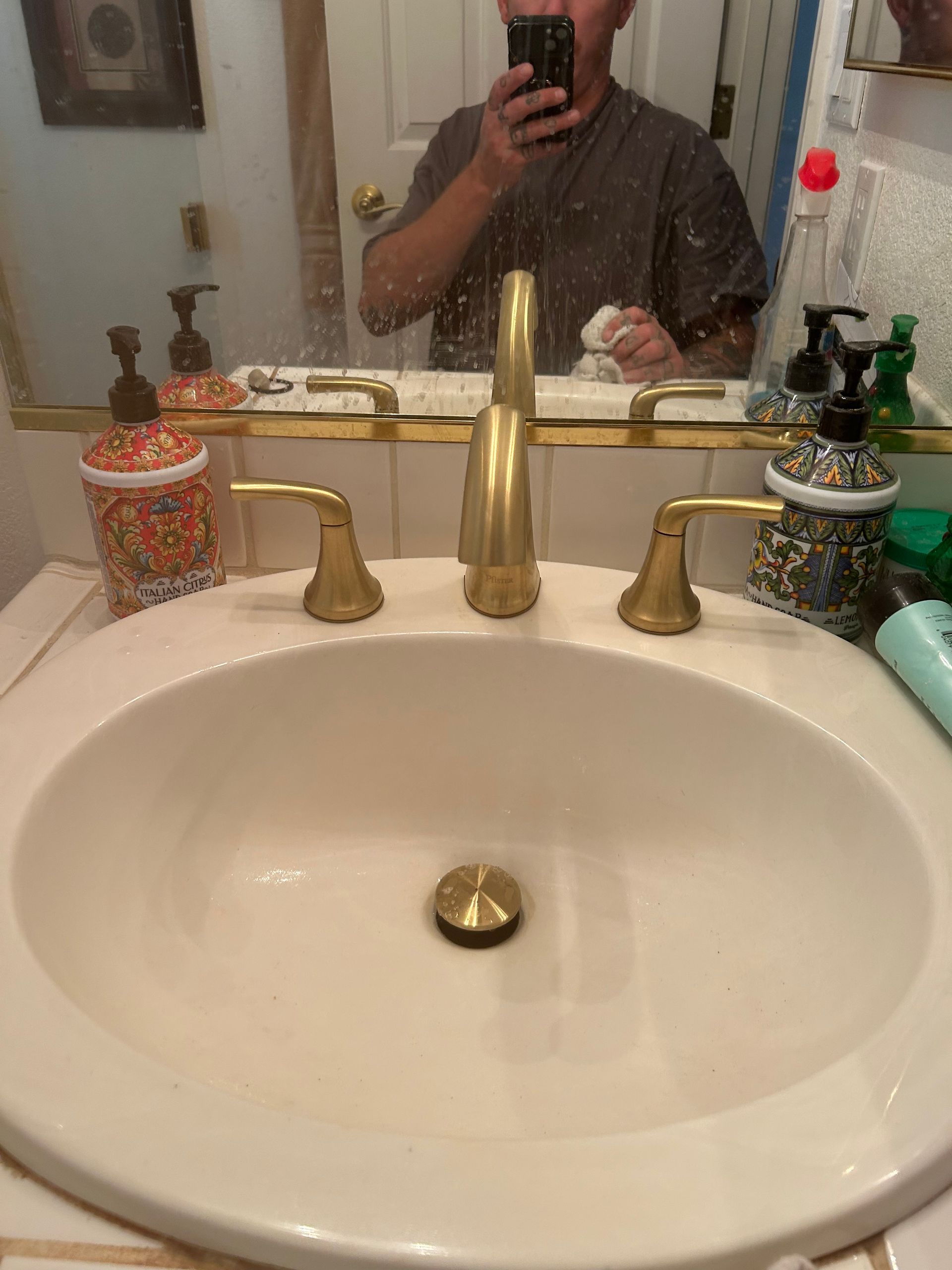 Bathroom sink with gold faucet and mirror, showing a person taking a photo in the reflection.