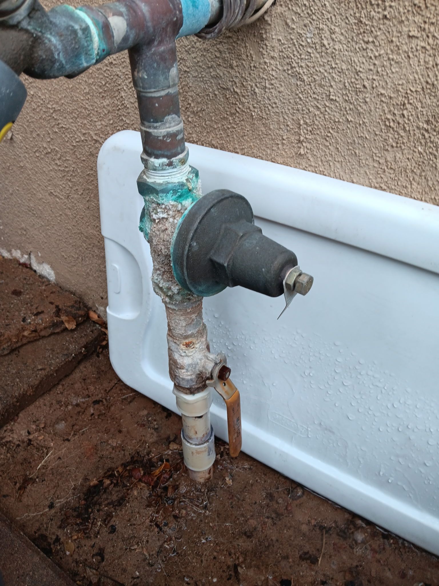 Corroded outdoor pipe connected to a white tank on muddy ground.