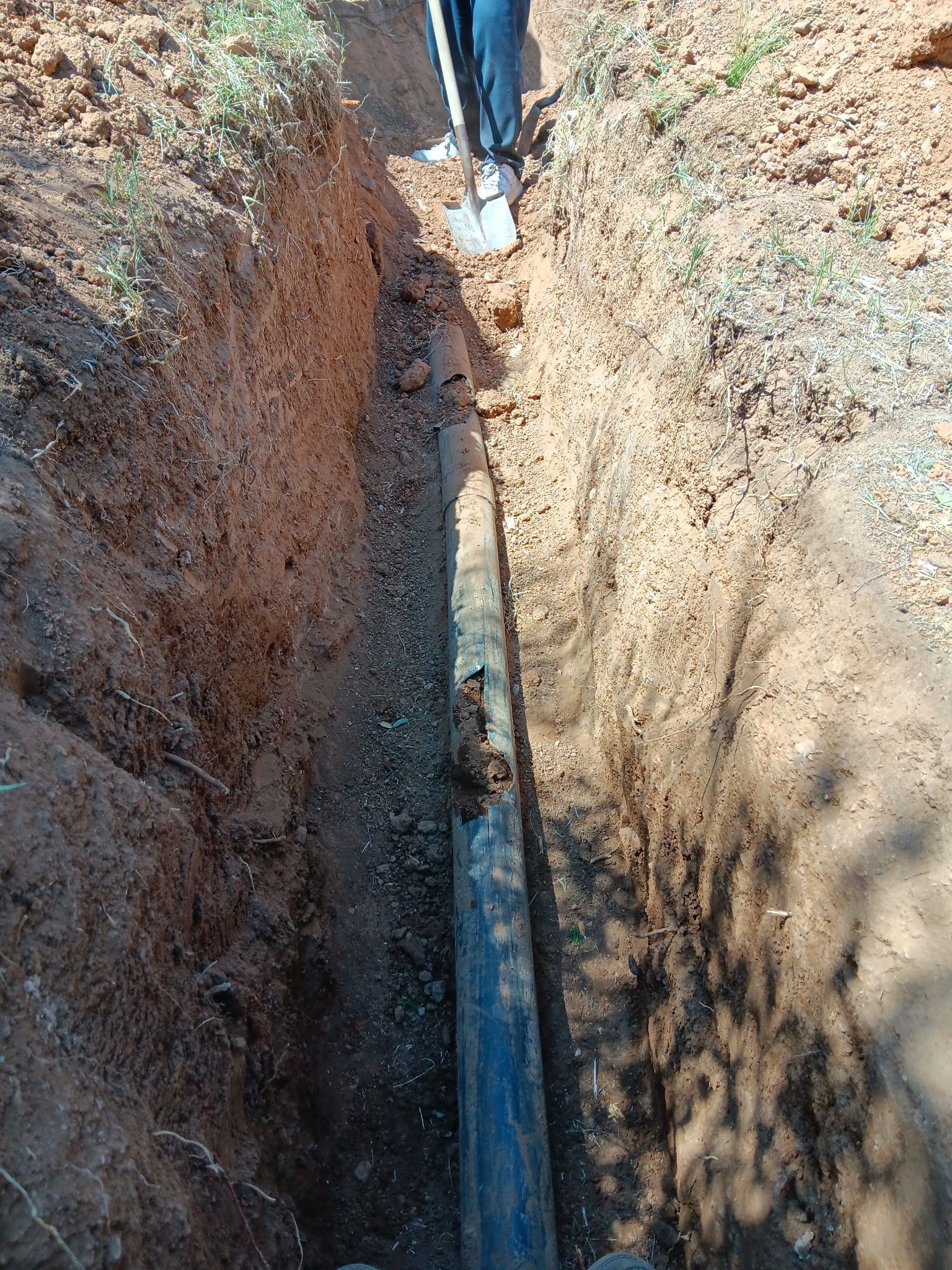 Trench with a black pipe laid in the ground and a shovel at the top end.