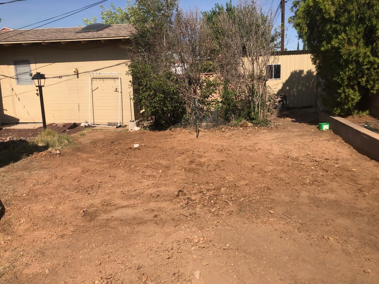 Dry dirt yard with a beige garage on the left and trees and a small building in the background.