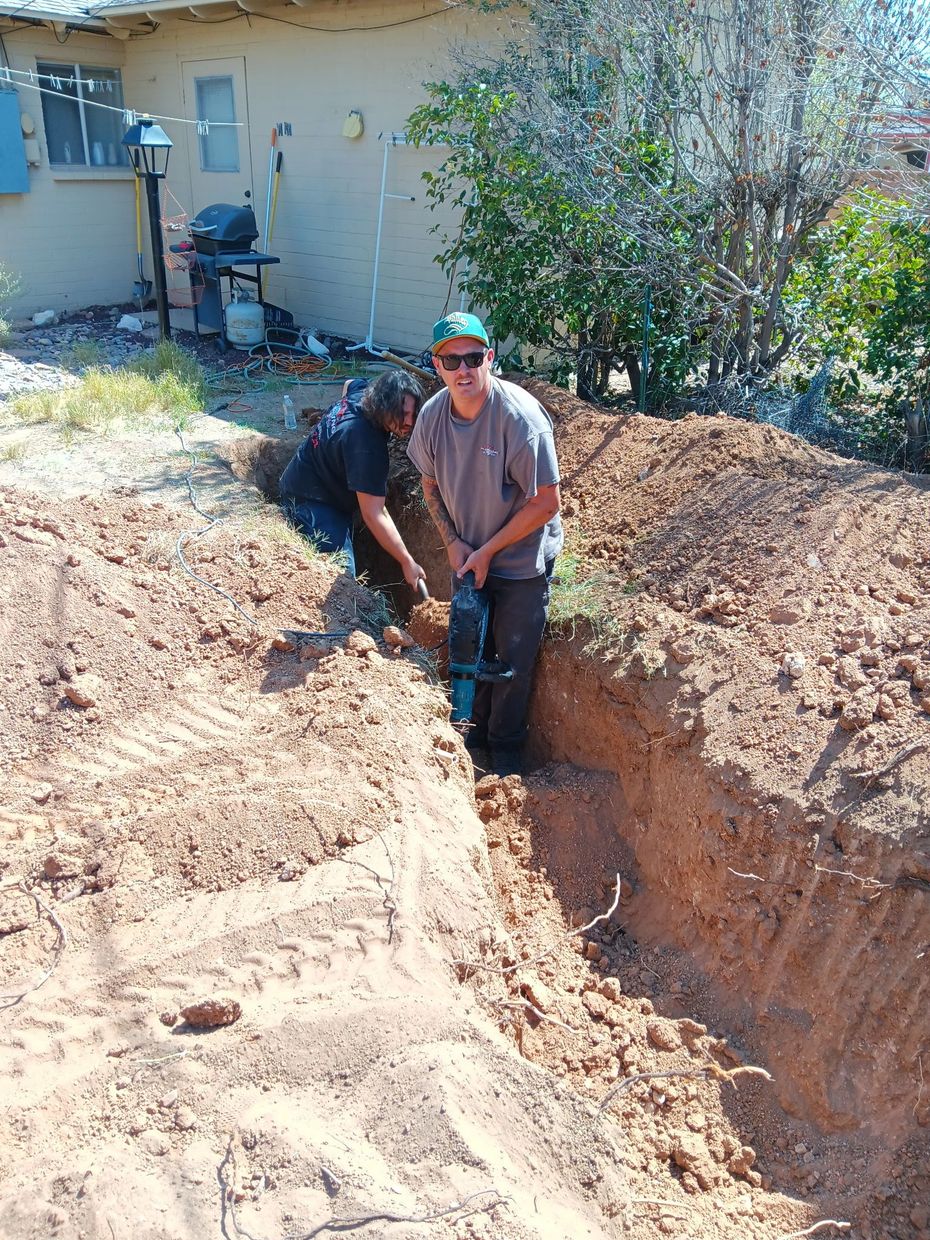 Two workers standing in a deep trench beside a house, surrounded by piles of dirt