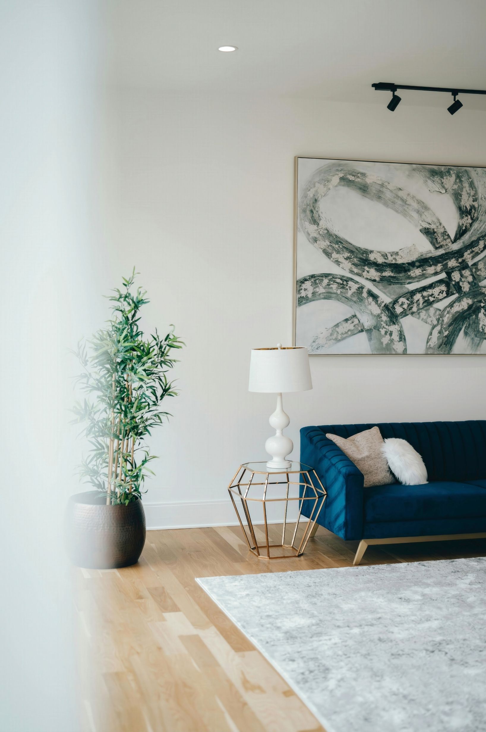 Modern living room with blue sofa, floor lamp, tall plant, abstract wall art, and light wood floors.