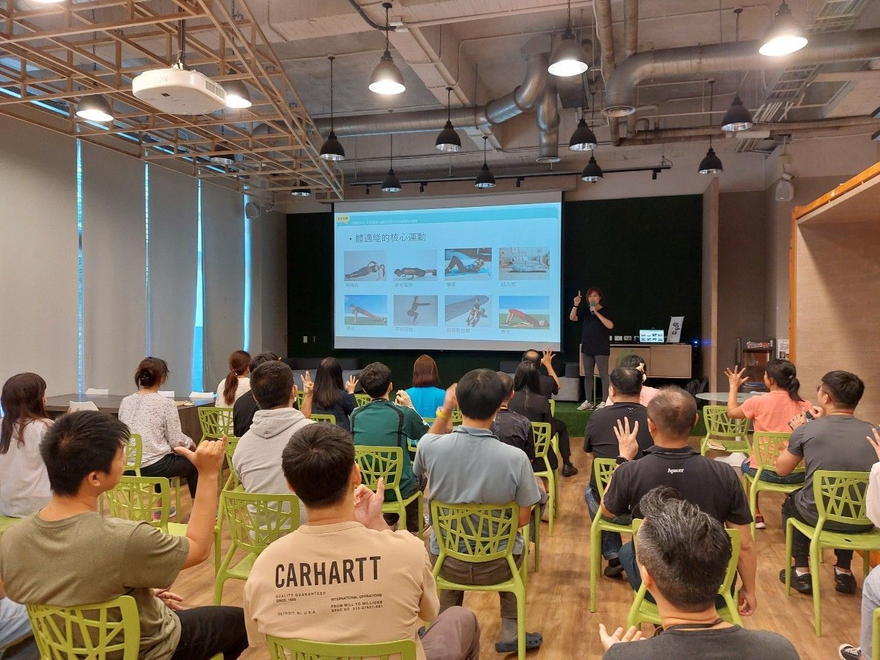 一位演讲者在一个明亮的会议室里向听众讲话,会议室里有绿色的椅子和一个播放幻灯片的屏幕。