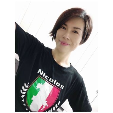 一位面带微笑、留着深色短发的人穿着一件黑色T恤，上面印有意大利国旗盾形标志，并标有“Nicolas”字样。
