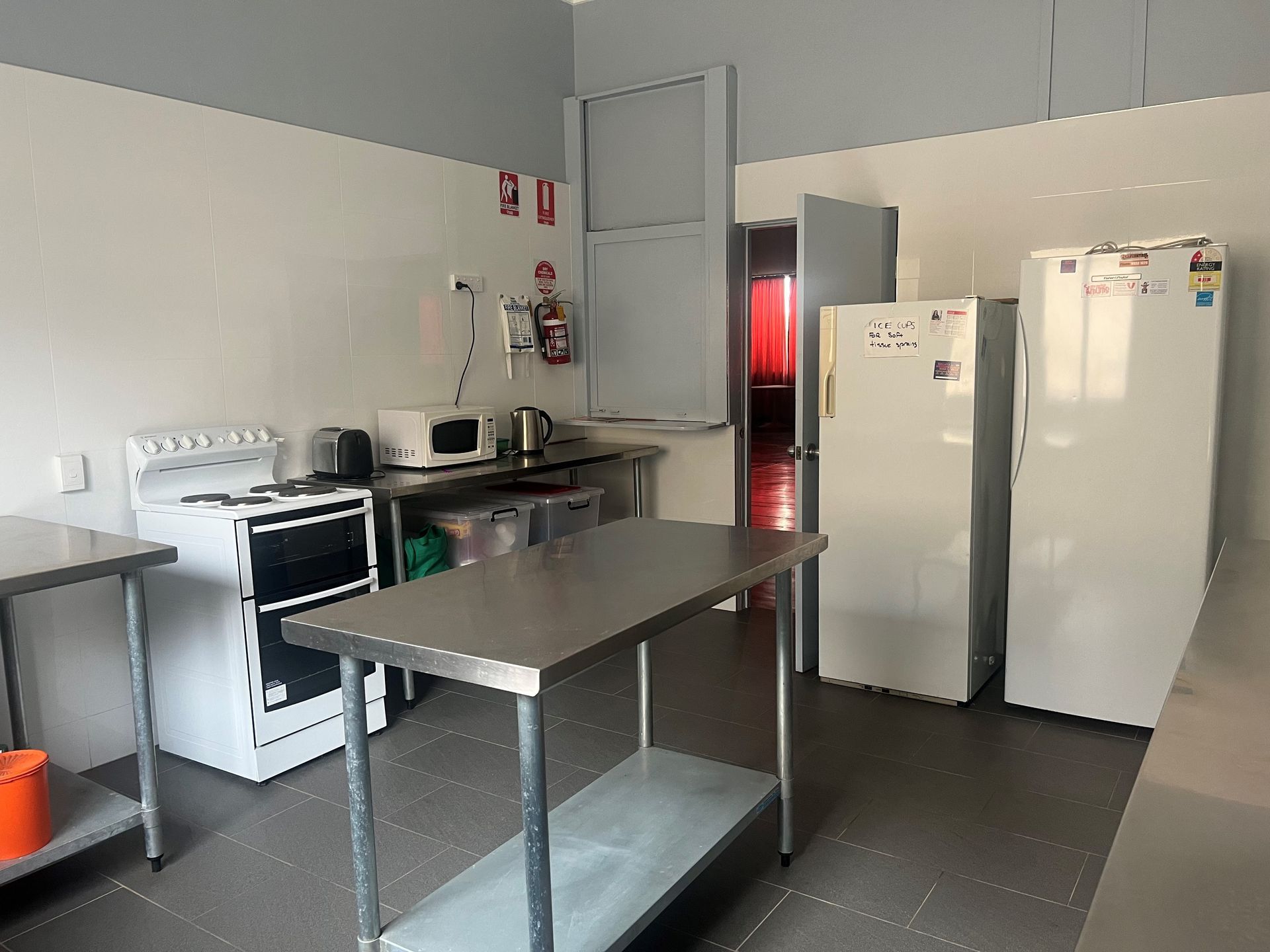 Kitchen with stainless steel prep table, appliances, and two refrigerators. White walls, grey floor.
