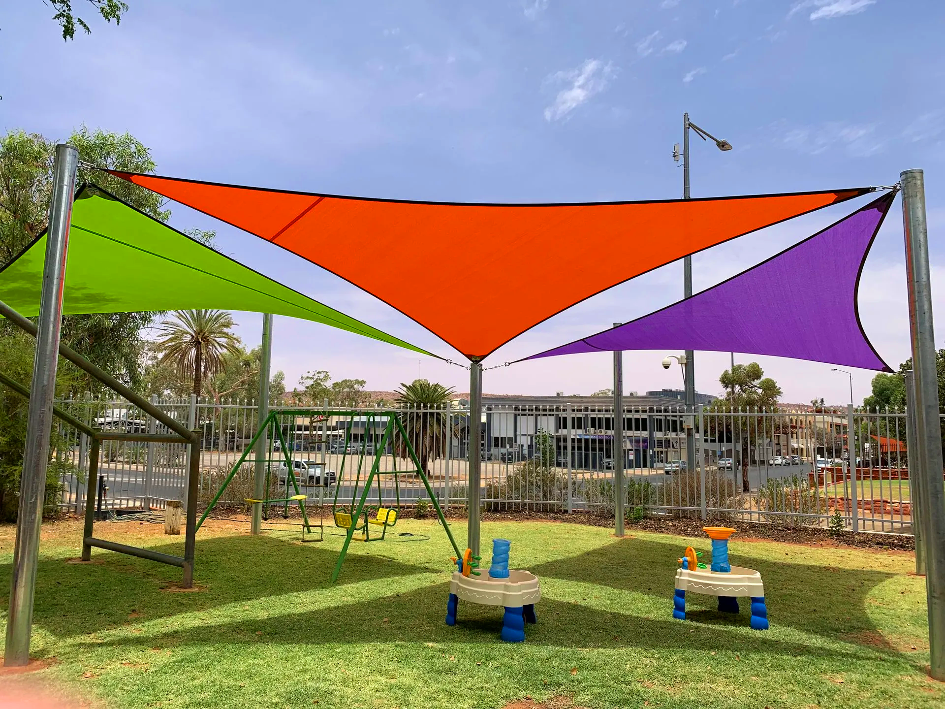 Colorful shade sails over a playground with swings and play tables, under a blue sky.