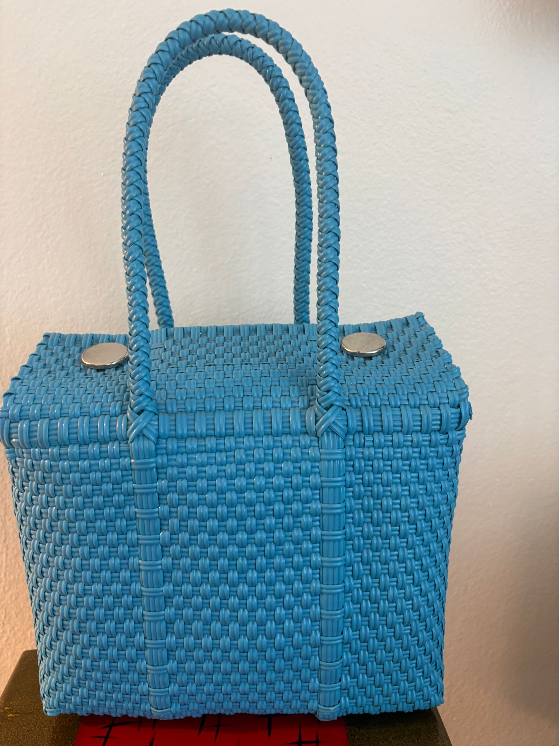 Blue woven tote bag with double handles and silver snaps, standing against a beige wall