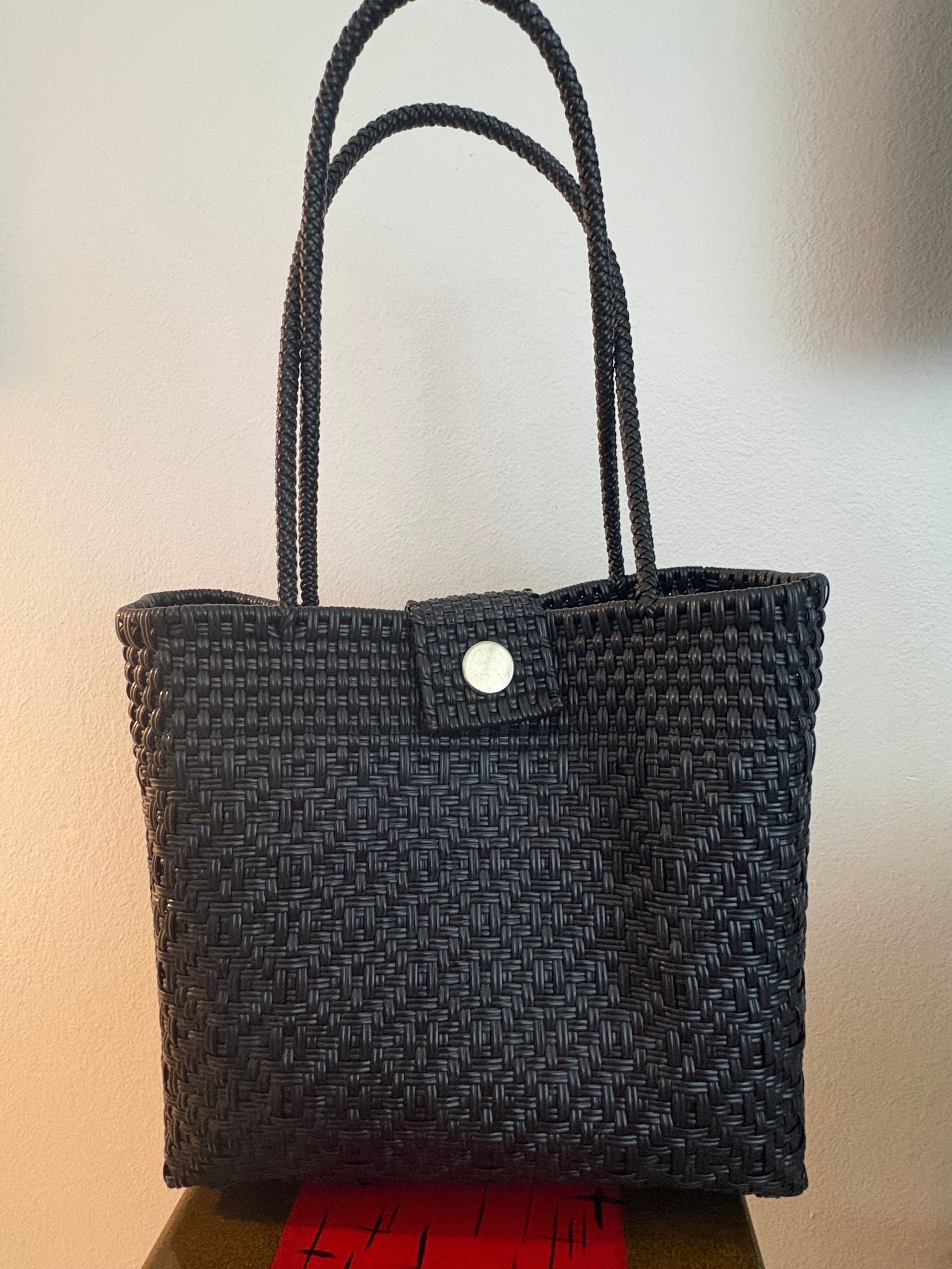 Black woven tote bag with long handles and a white button clasp against a light wall.