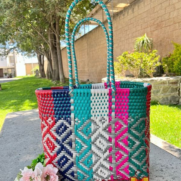 Colorful woven tote bag with turquoise handles, standing on a sidewalk beside a grassy yard and stone wall