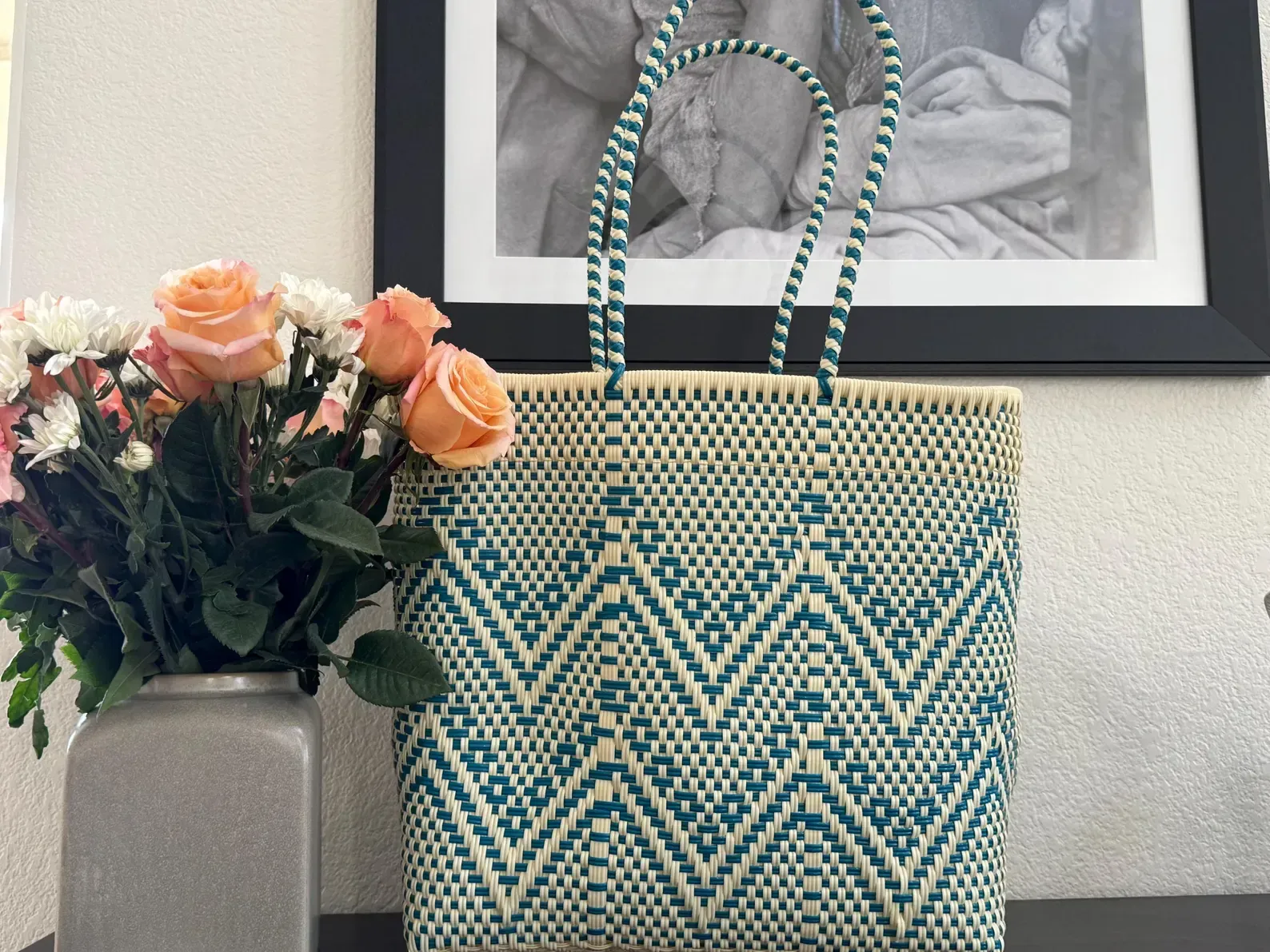 Blue and cream woven tote bag beside a vase of peach flowers under a framed black-and-white photo