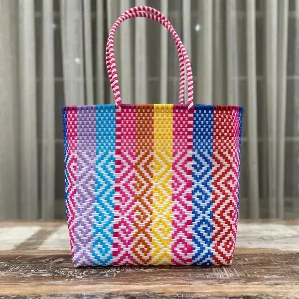 Colorful woven tote bag with pink handles on a wooden surface in front of gray curtains