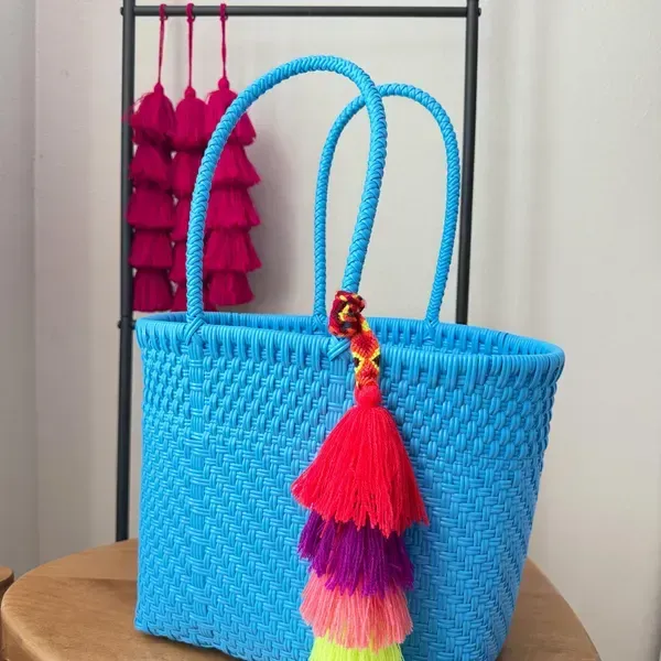 Bright blue woven tote bag with colorful tassel on a table, with pink hanging tassels in the background