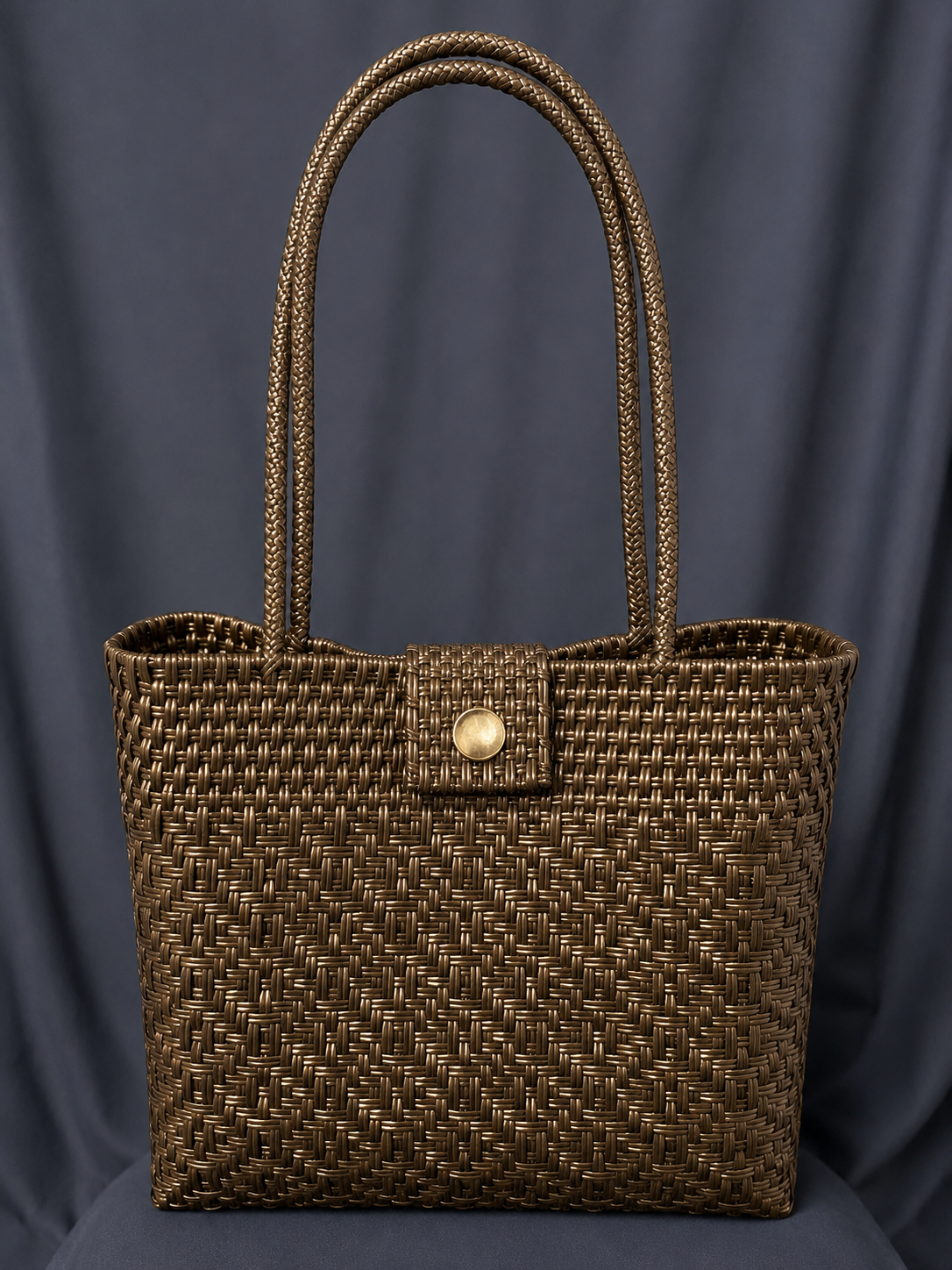 Brown woven handbag with tall handles and a gold clasp, displayed against a dark gray background