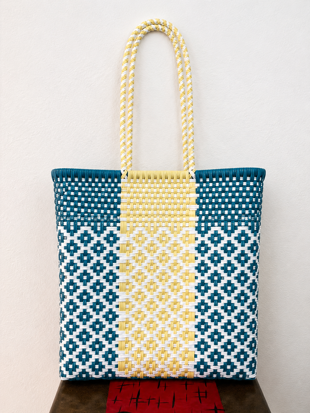 Woven tote bag with blue and cream pattern and yellow handles, standing on a red base.