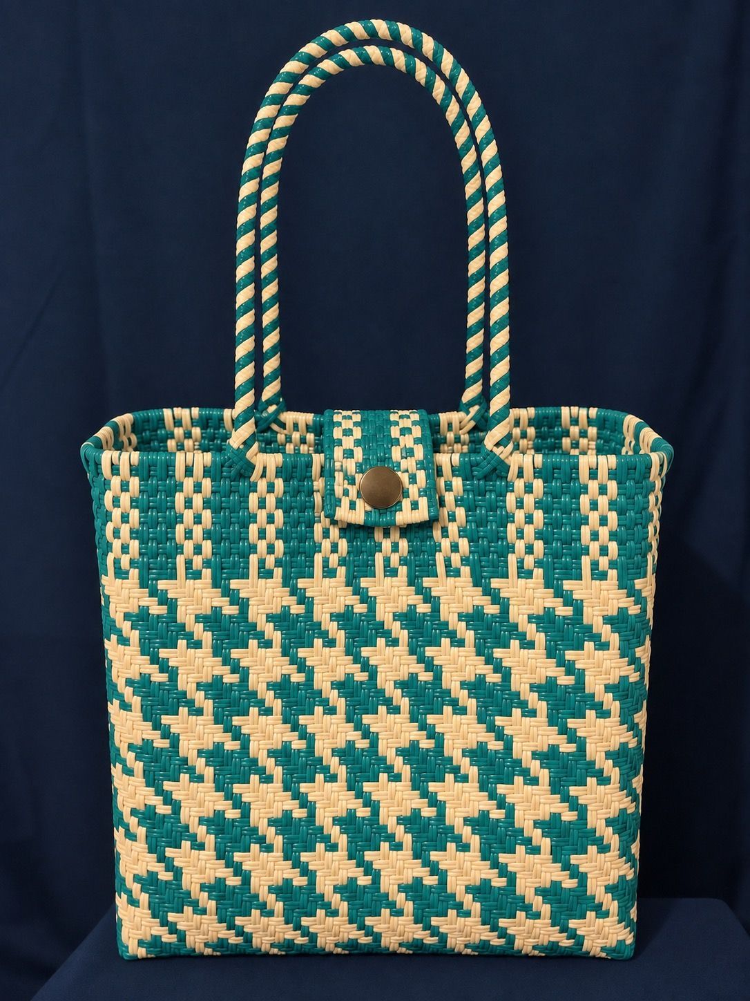 Teal and beige woven tote bag with braided handles and a buckle clasp on a dark background