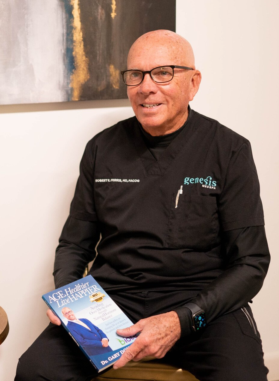 A bald man is sitting on a stool holding a book.