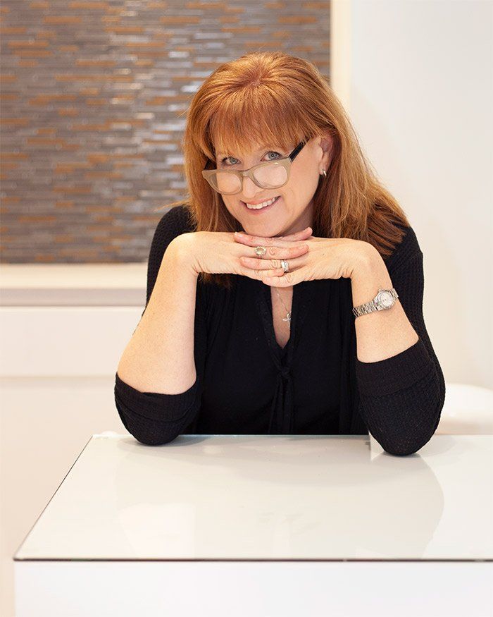 A woman wearing glasses is sitting at a table with her hands on her chin.