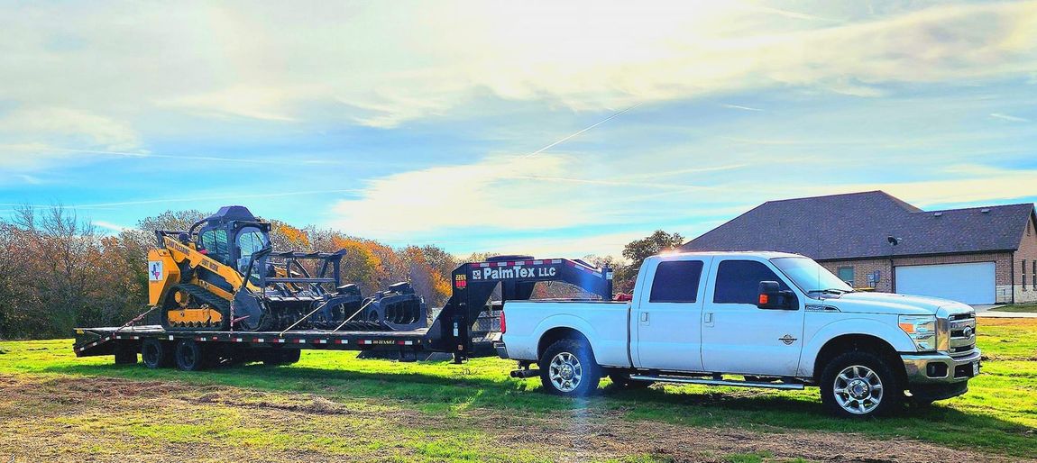 A white pickup truck pulling a flatbed trailer loaded with a yellow tracked skid steer in a grassy field.