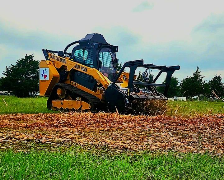 A yellow skid steer with a forestry mulcher attachment clearing brush in a grassy field under a cloudy sky.