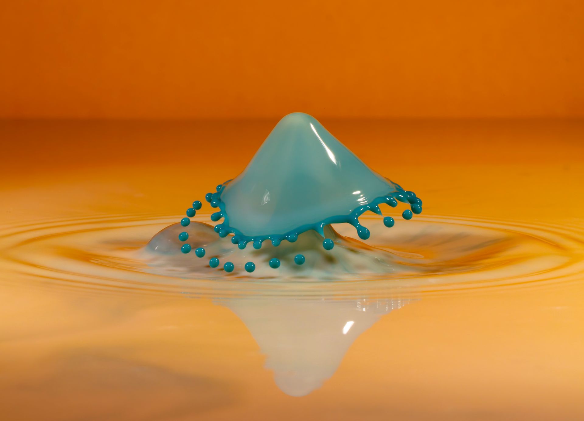 Turquoise water droplet crown emerging from milky liquid on a yellow-orange surface.