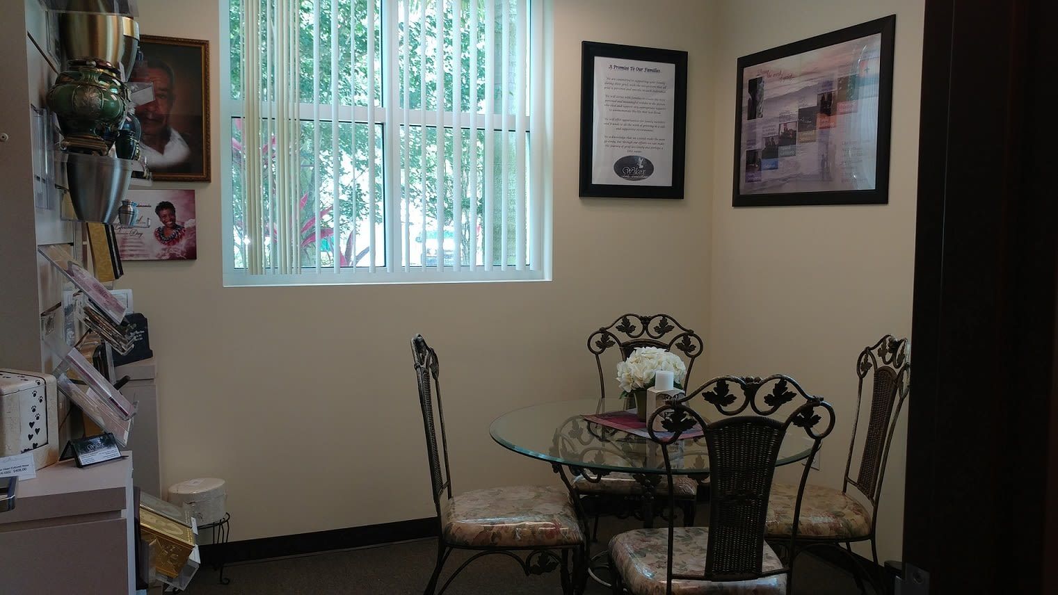 Small dining area with glass table and chairs, framed wall art, and a window with vertical blinds.