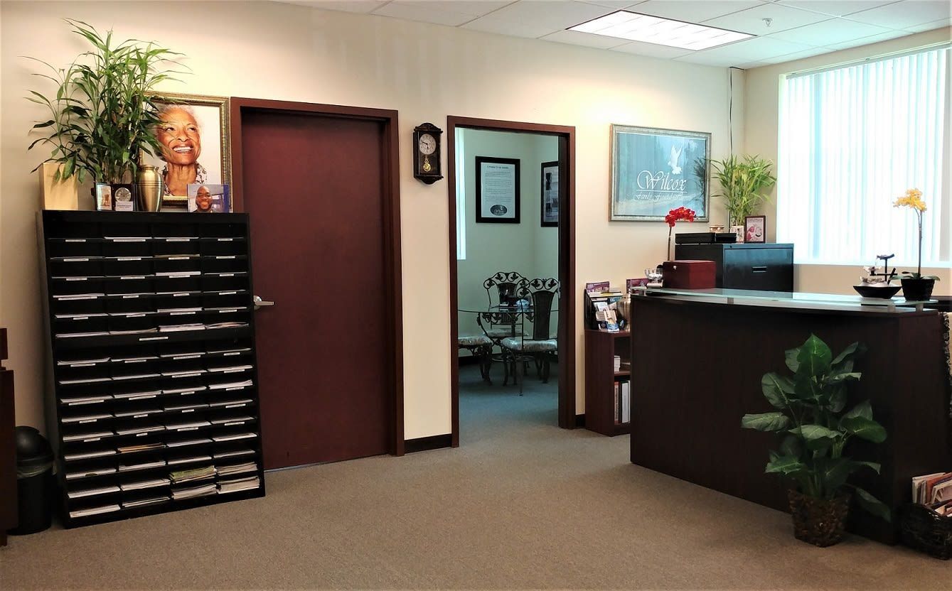 Office reception area with dark wood desk, mail cubbies, potted plants, and open doorway to a back room