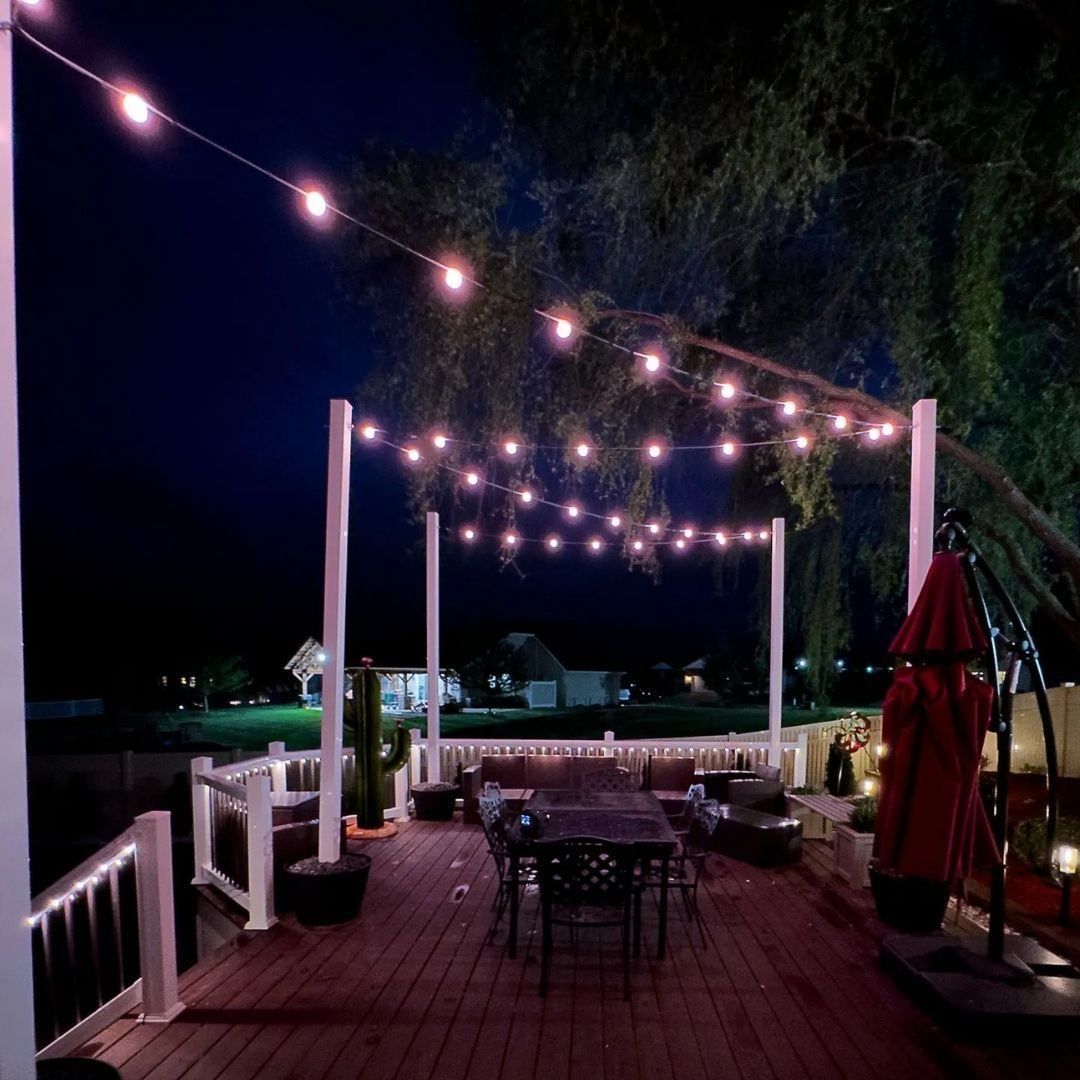 String lights over a wooden deck dining area at night beside a pool and umbrellas