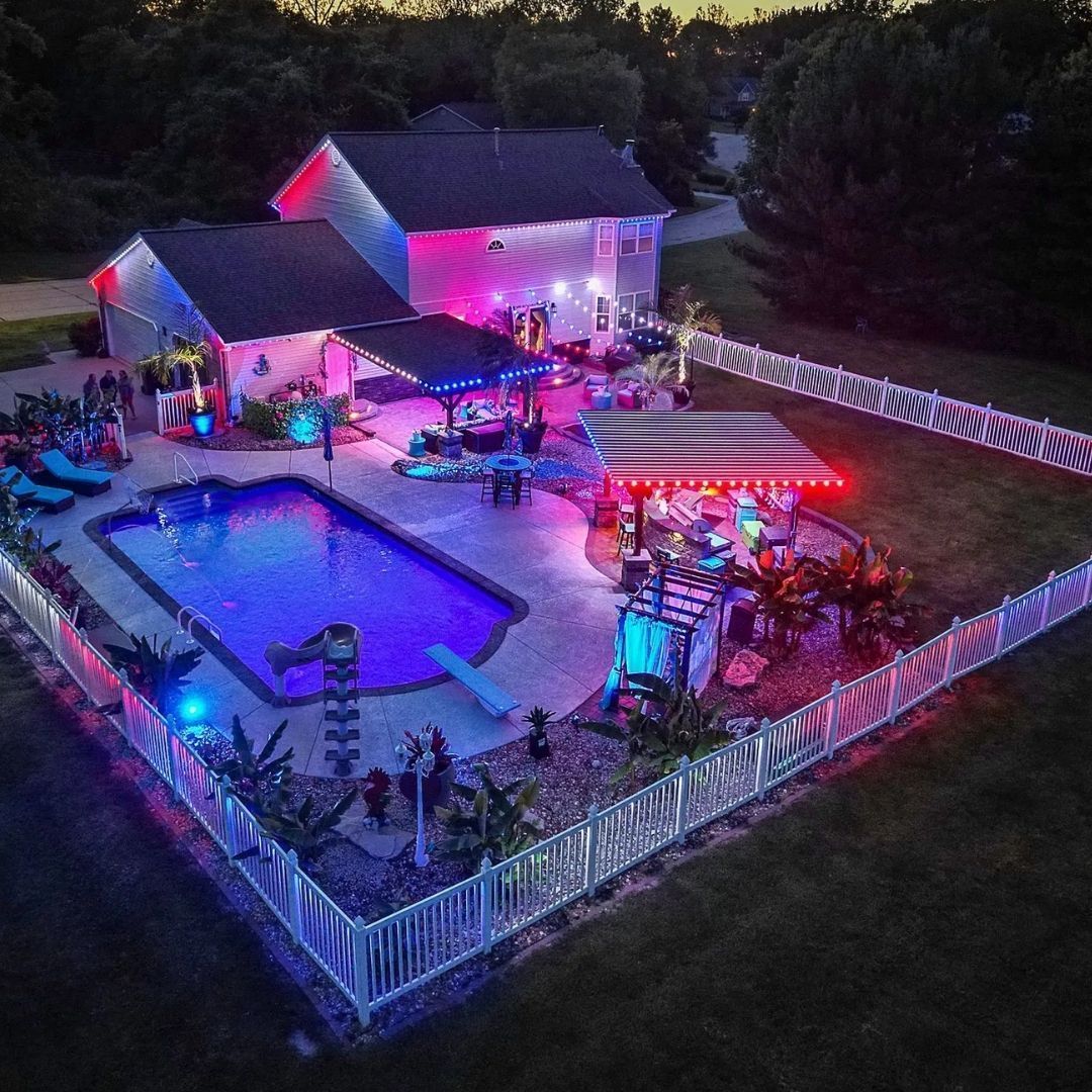 Colorful lit backyard patio with a pool, pergola seating, and a house at dusk, surrounded by white fencing