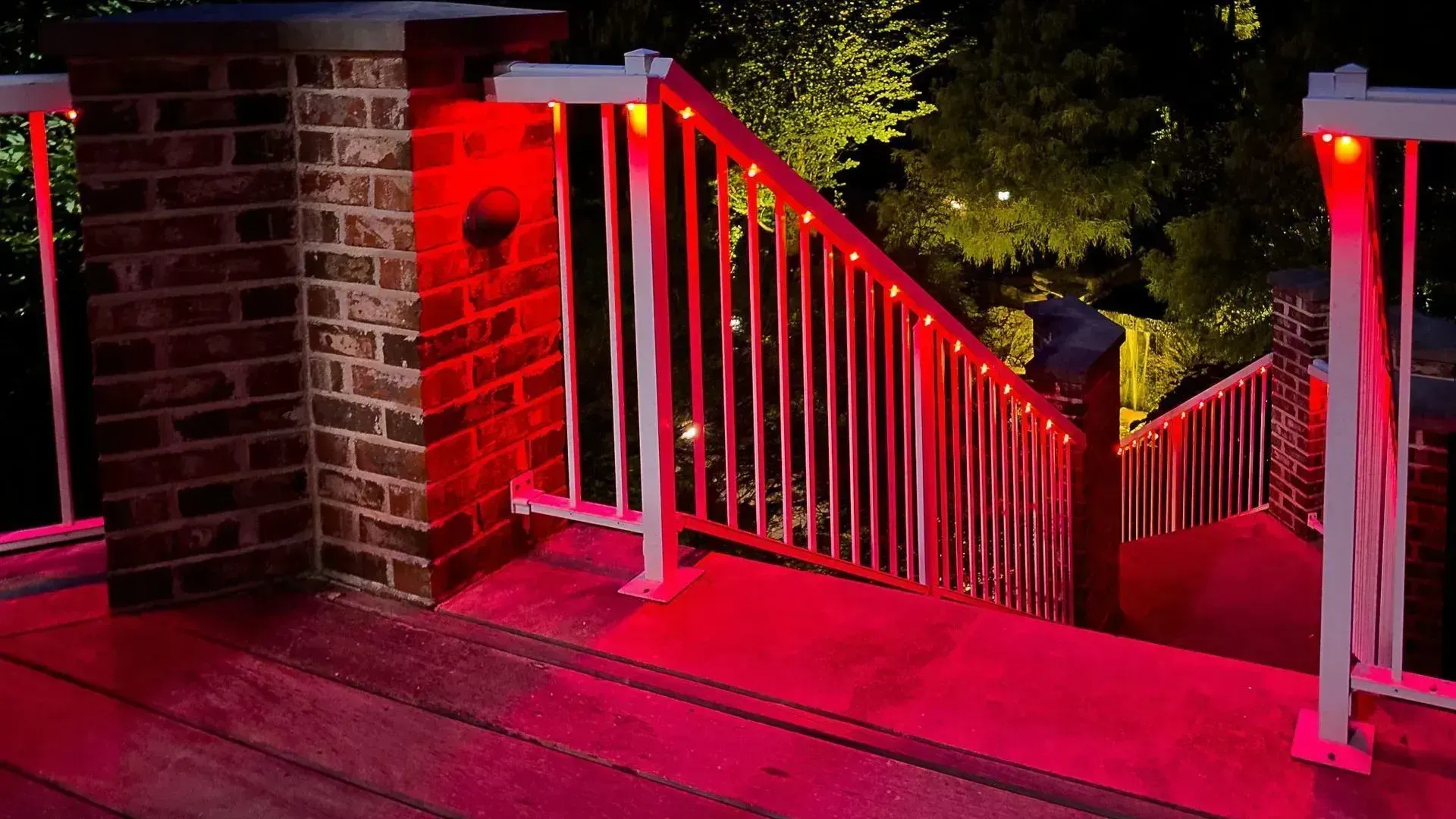 Red-lit outdoor wooden stairs and deck with white railings beside a brick pillar at night
