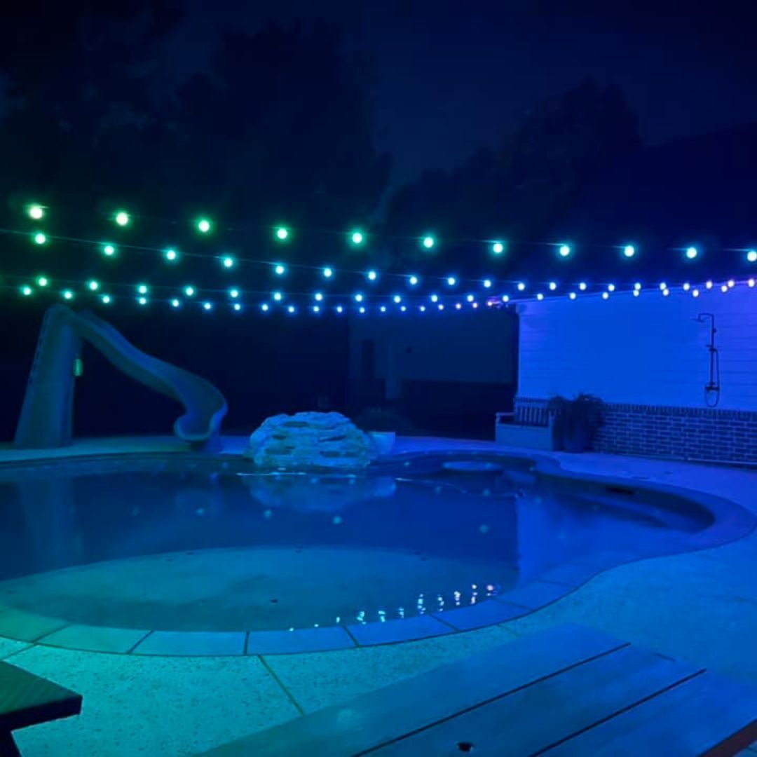 Blue-lit backyard pool at night with string lights overhead and patio chairs in the background