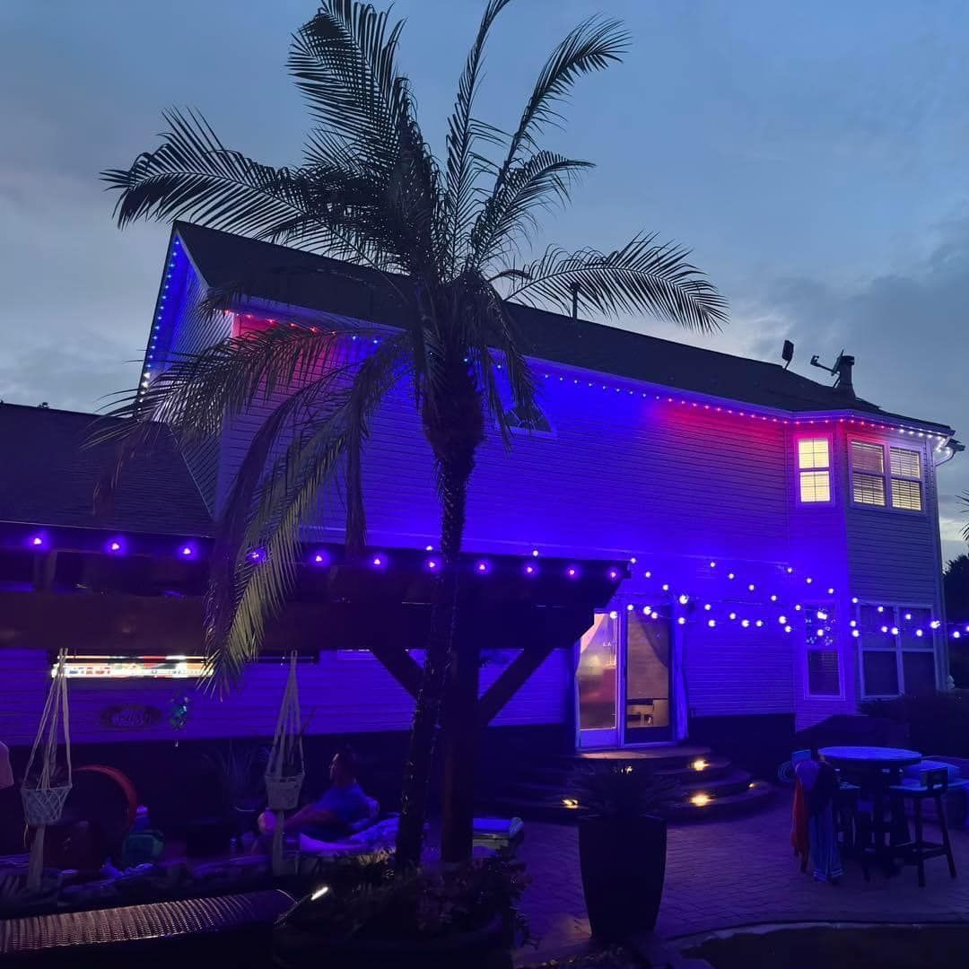 House with purple-blue string lights at dusk beside a palm tree and outdoor seating