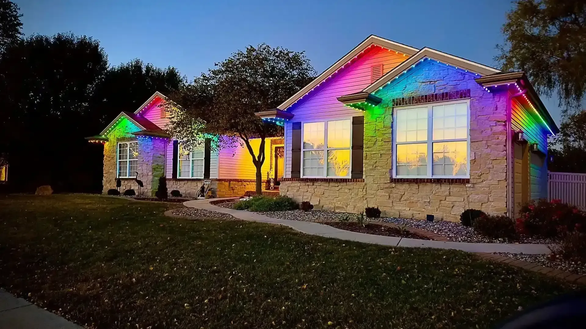 Two lit houses at dusk with rainbow-colored roof and window lights along a walkway