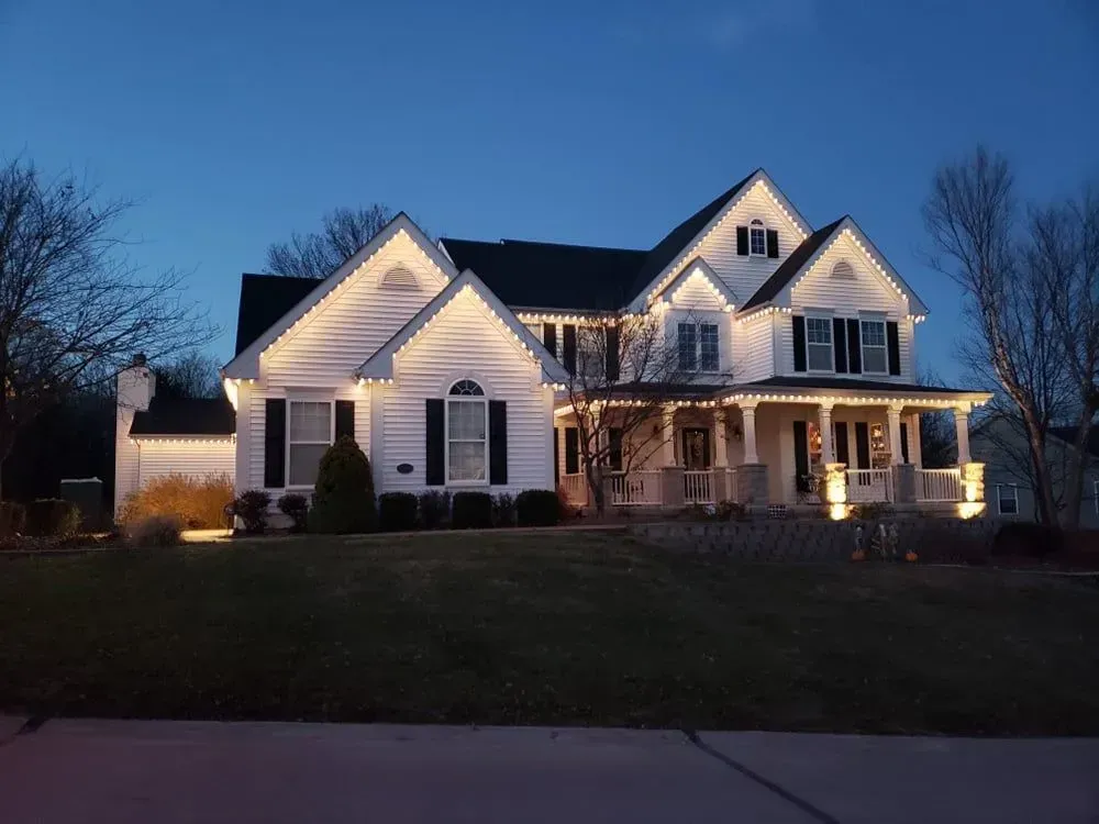 Illuminated white two-story suburban house at dusk with porch lights and a dark lawn