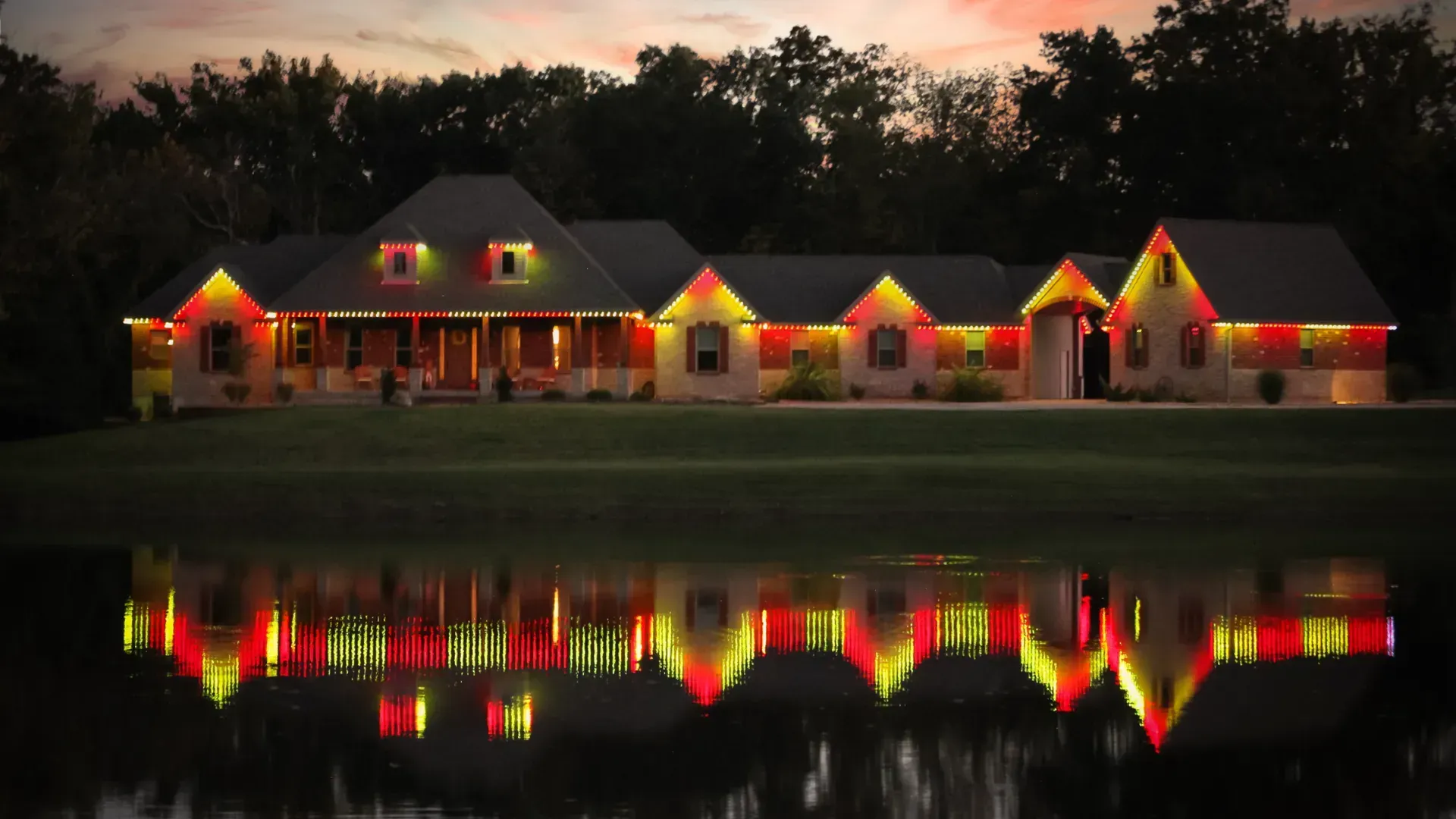 A lit house with red and yellow lights reflected in a calm lake at dusk