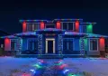 Two-story house decorated with colorful Christmas lights at night, with snow on the ground.