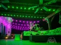Green and purple-lit outdoor patio with sofas, string lights, and a ceiling fan at night