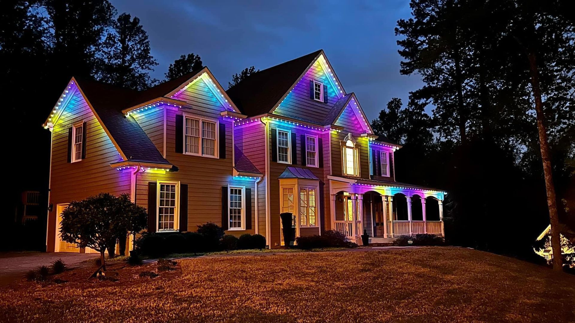 Two-story house outlined in colorful holiday lights at night in a wooded yard