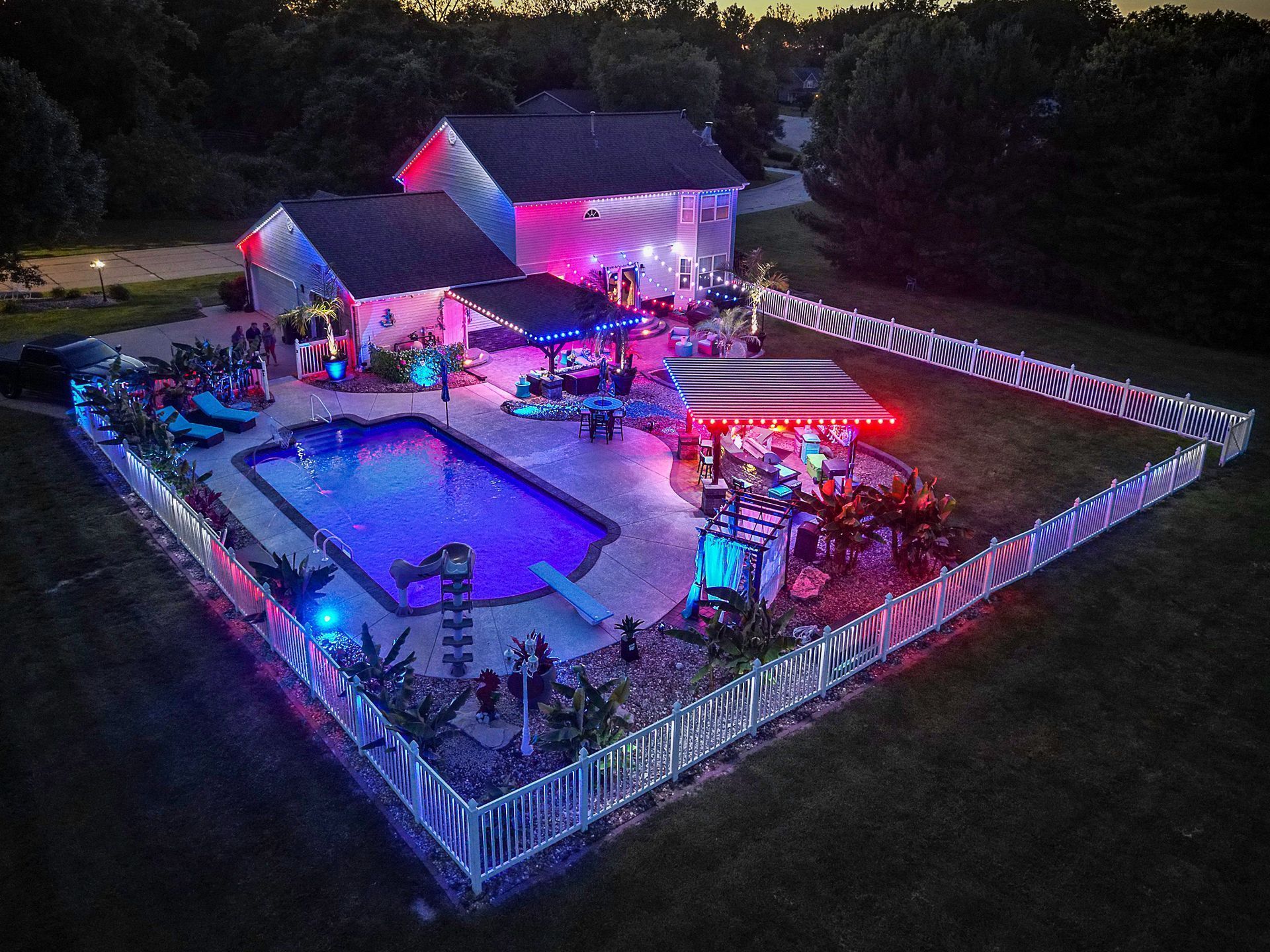 Nighttime outdoor pool party with colorful lights, tents, and guests in a fenced backyard