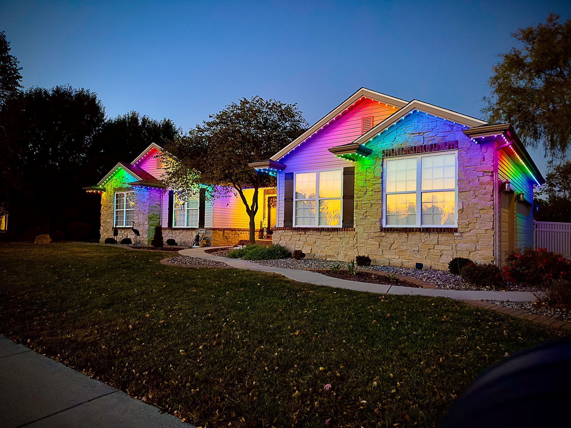 Single-story house at dusk with rainbow-colored Christmas lights along the roofline and windows.