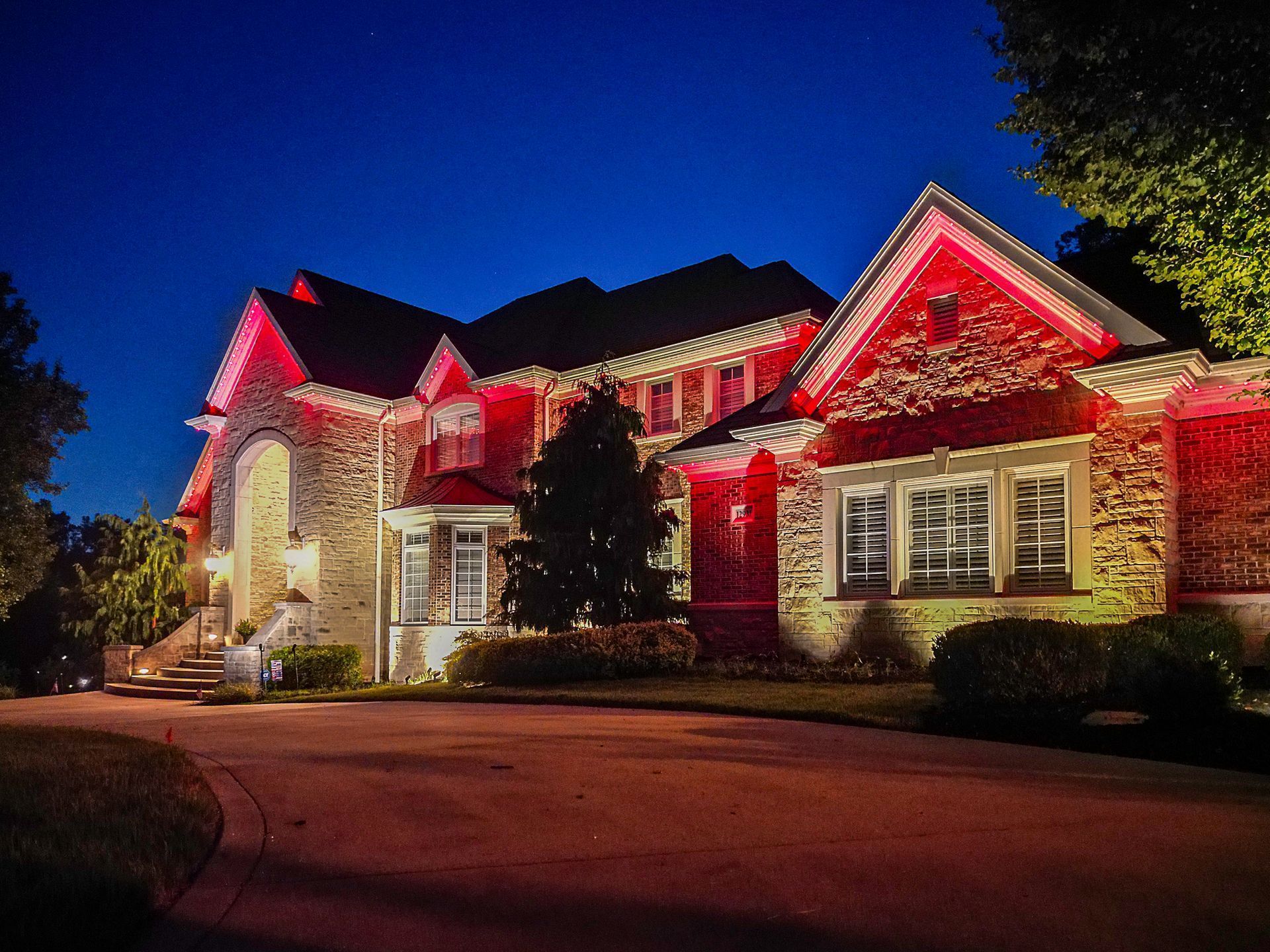 Large brick house lit with red holiday lights at dusk, viewed from the driveway